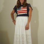 Woman wearing a colorful top with an American flag design and a white skirt against a plain background