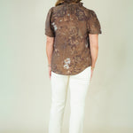 A woman viewed from behind wearing a brown short-sleeve button-up shirt with a subtle floral pattern, paired with white pants and beige shoes, against a plain light beige studio background.