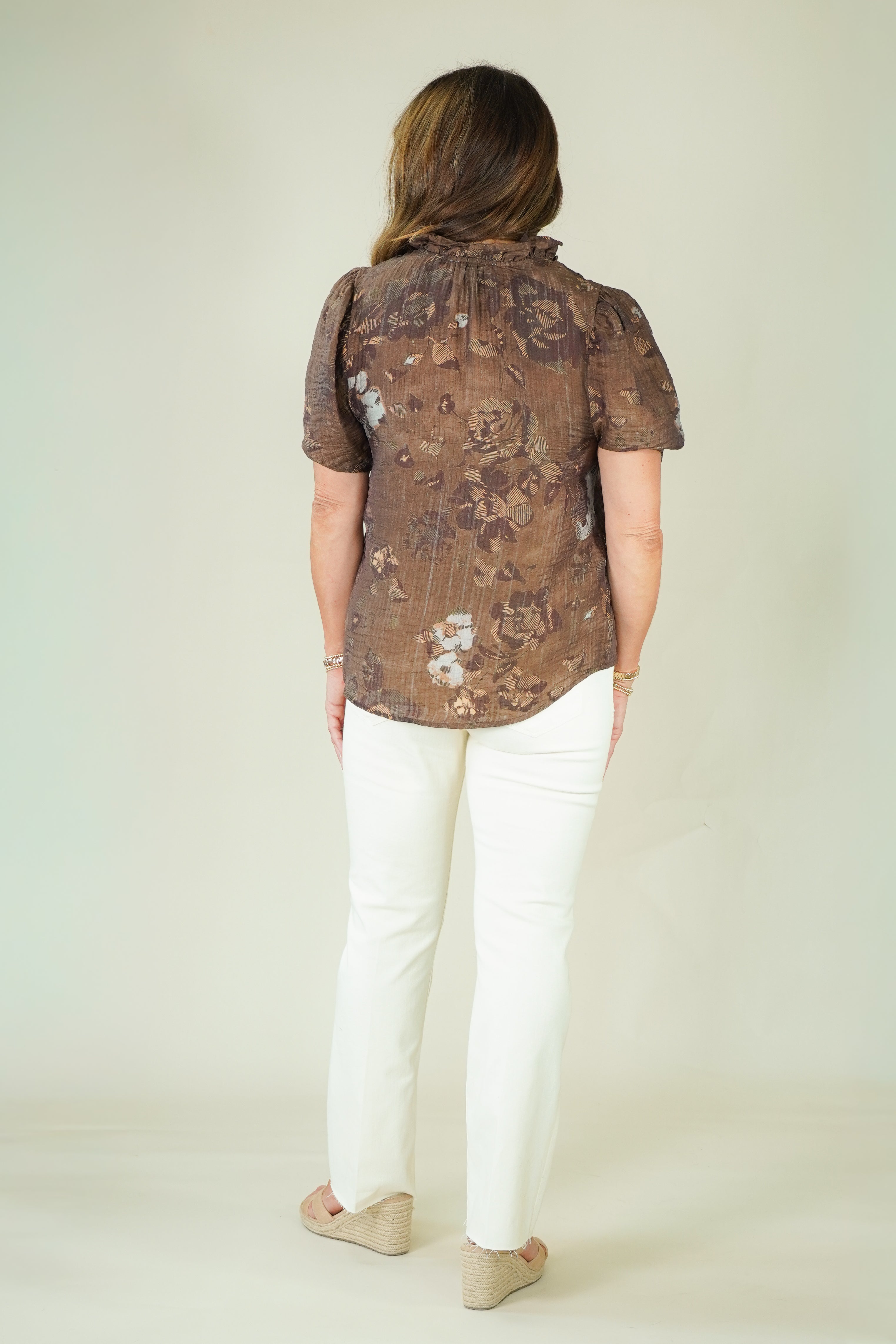 A woman viewed from behind wearing a brown short-sleeve button-up shirt with a subtle floral pattern, paired with white pants and beige shoes, against a plain light beige studio background.