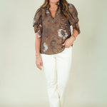 A woman modeling a brown and beige abstract patterned blouse with puff sleeves, paired with white pants and beige sandals, standing against a plain light beige studio background.