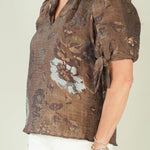 A woman modeling a short-sleeve brown patterned blouse with white pants and beige wedge sandals, standing against a plain light beige studio background.