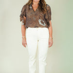A woman modeling a brown and beige floral patterned blouse with short puff sleeves, paired with white pants and beige wedge sandals, standing against a light beige studio background.