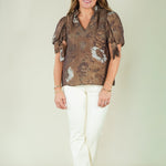 A woman modeling a brown short-sleeve blouse with a white abstract pattern and white pants, standing against a plain light beige studio background.