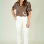 A woman modeling a brown patterned short-sleeve blouse with white straight-leg pants and beige wedge sandals, standing against a plain light beige studio background.