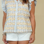 A woman wearing a white short-sleeve button-up blouse with a yellow and blue floral pattern, ruffled sleeves, and white shorts, standing against a solid beige background.