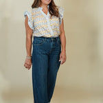 A woman modeling a light blue and yellow patterned short-sleeve blouse with ruffled shoulders, paired with dark blue wide-leg jeans and beige wedge sandals, against a solid beige studio background.