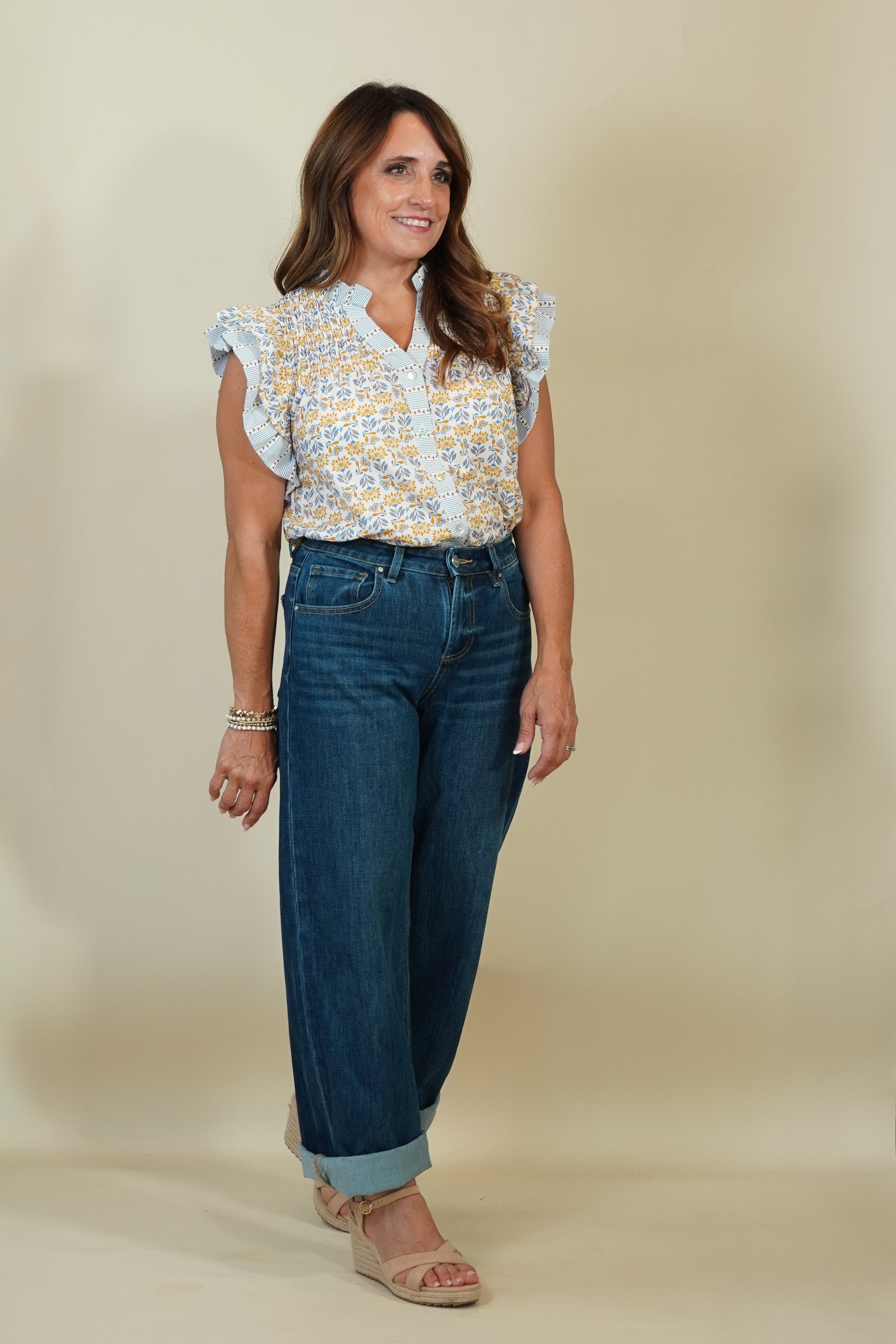 A woman modeling a light blue and yellow patterned short-sleeve blouse with ruffled shoulders, paired with dark blue wide-leg jeans and beige wedge sandals, against a solid beige studio background.