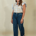 A woman modeling a white short-sleeve blouse with a yellow and blue patterned print and ruffled sleeves, paired with dark blue wide-leg jeans and beige wedge sandals, against a solid beige studio background.