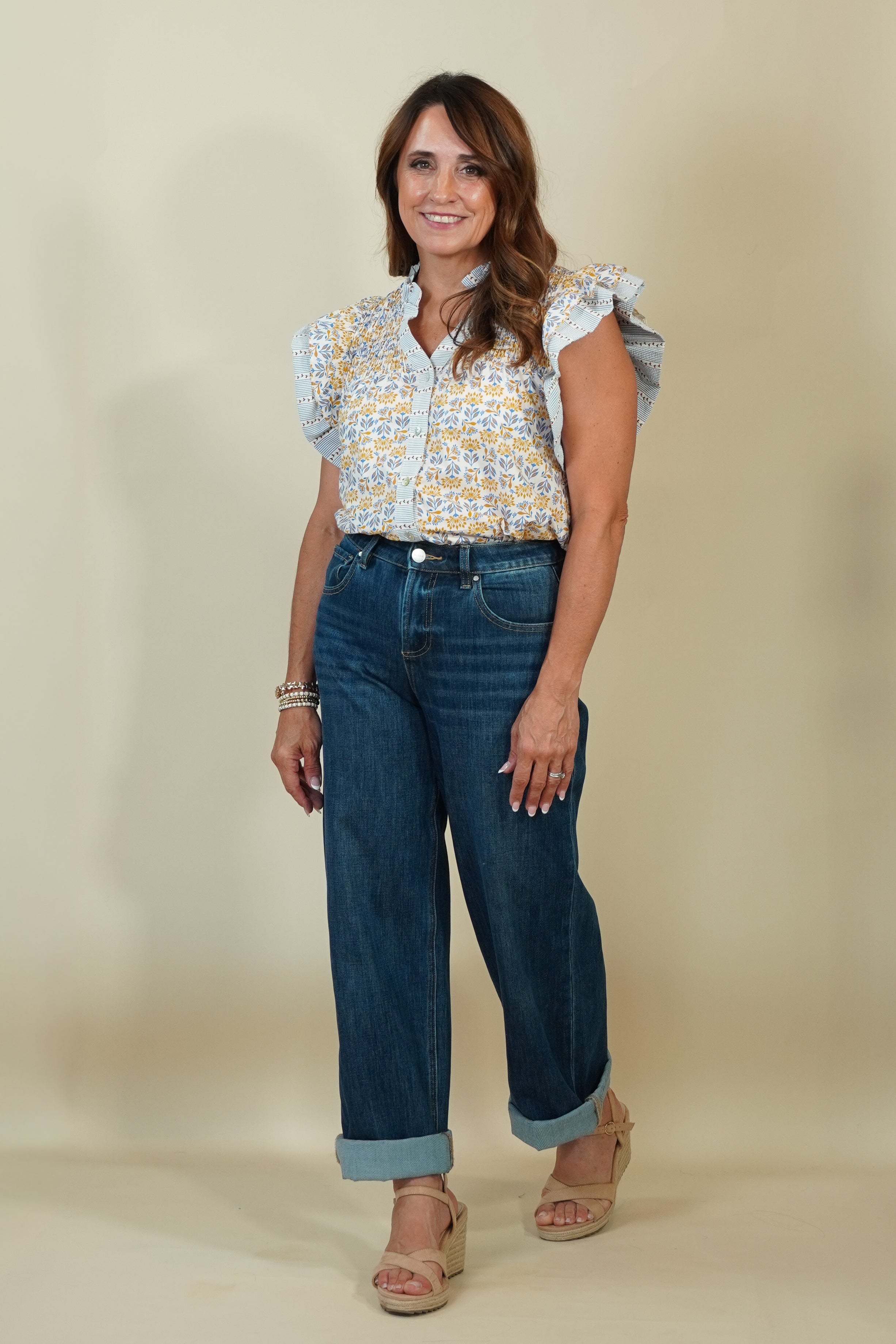 A woman modeling a white short-sleeve blouse with a yellow and blue patterned print and ruffled sleeves, paired with dark blue wide-leg jeans and beige wedge sandals, against a solid beige studio background.