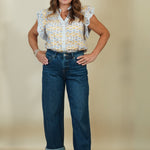 A woman modeling a sleeveless white blouse with a yellow and blue geometric pattern, paired with dark blue jeans and beige wedge sandals, against a solid beige studio background.