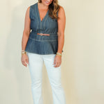 A woman modeling a sleeveless blue denim peplum top with a thin tan belt, paired with white cropped pants and beige wedge sandals, against a plain cream background.