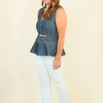 A woman modeling a sleeveless blue denim vest with a peplum hem and a thin brown belt, paired with white cropped pants and beige wedge sandals, against a plain cream background.