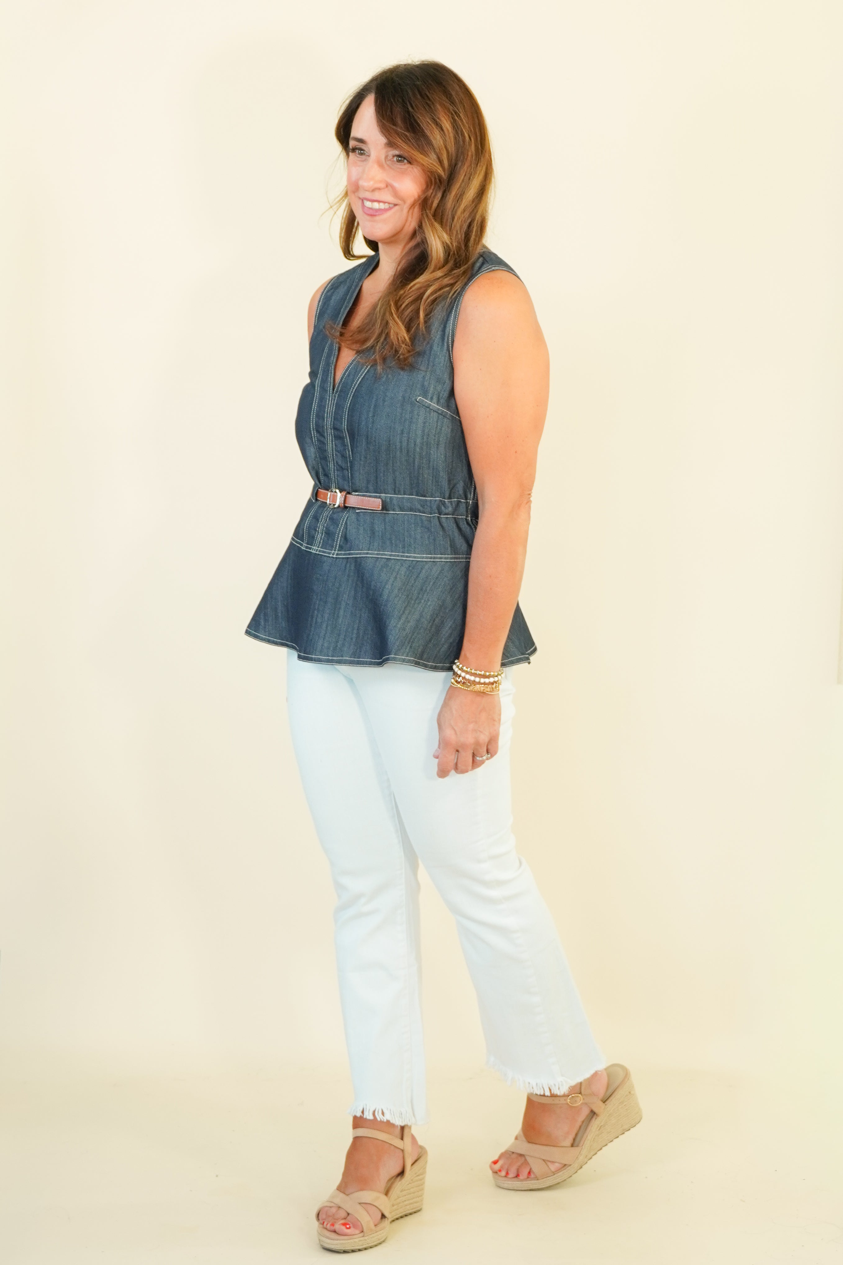 A woman modeling a sleeveless blue denim vest with a peplum hem and a thin brown belt, paired with white cropped pants and beige wedge sandals, against a plain cream background.
