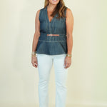 A woman modeling a sleeveless denim vest with a brown belt, paired with white cropped pants and beige wedge sandals, standing against a plain cream background.