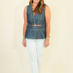 A woman modeling a sleeveless blue denim vest with a tan belt, paired with white pants and tan sandals, standing against a plain cream background.