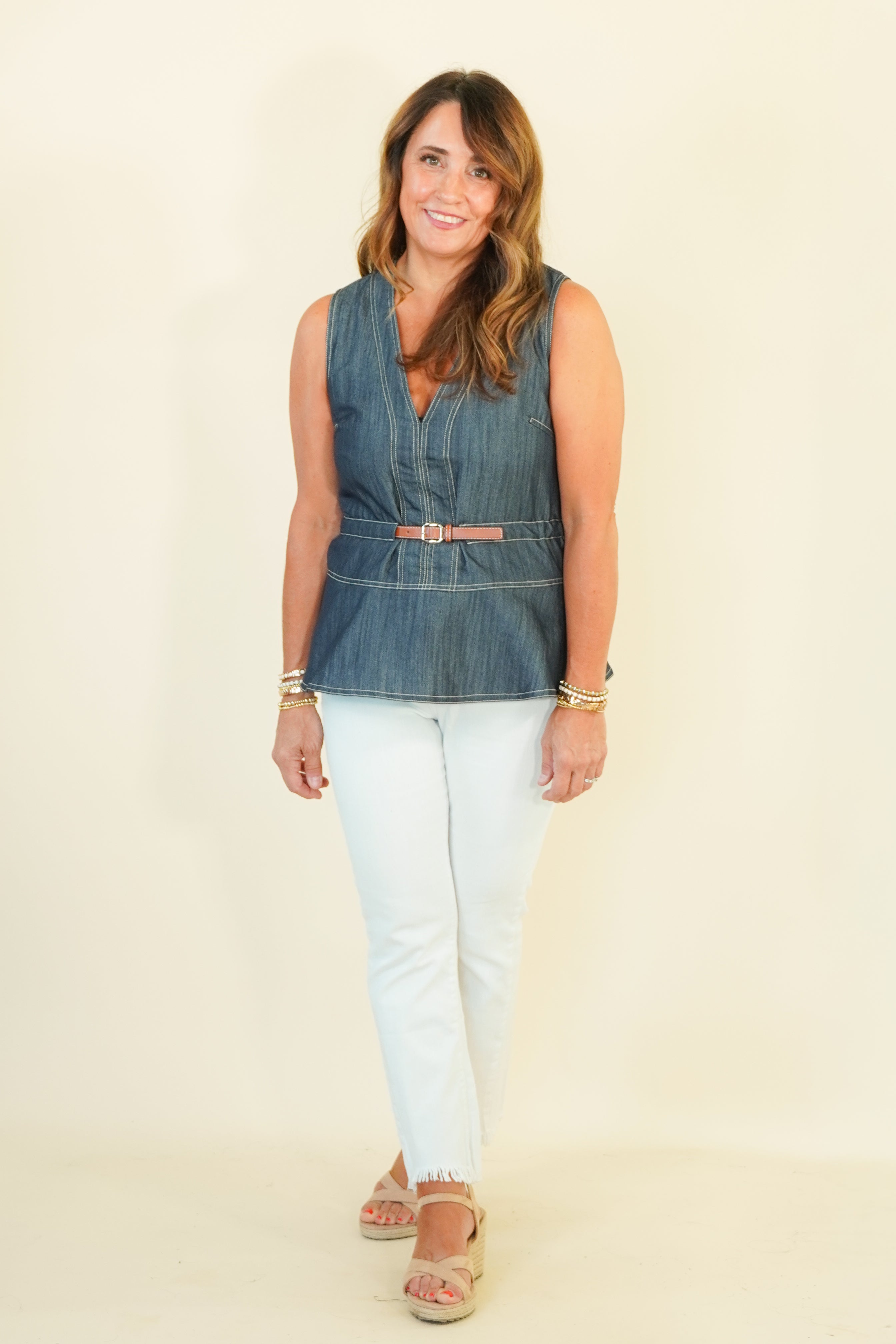 A woman modeling a sleeveless blue denim vest with a tan belt, paired with white pants and tan sandals, standing against a plain cream background.