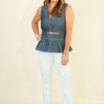 A woman modeling a sleeveless denim vest with a pink belt, paired with light-wash white pants and beige wedge sandals, standing against a solid cream background.