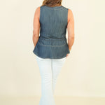 A woman viewed from the back wearing a sleeveless blue denim peplum top, white flared pants, and beige wedge sandals against a plain cream studio background.