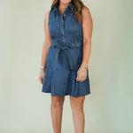 A woman modeling a sleeveless blue denim shirt dress with a matching tie belt, paired with beige wedge sandals, against a plain light green studio background.