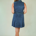 A woman viewed from behind wearing a sleeveless blue dress with a matching belt, standing against a plain light background.