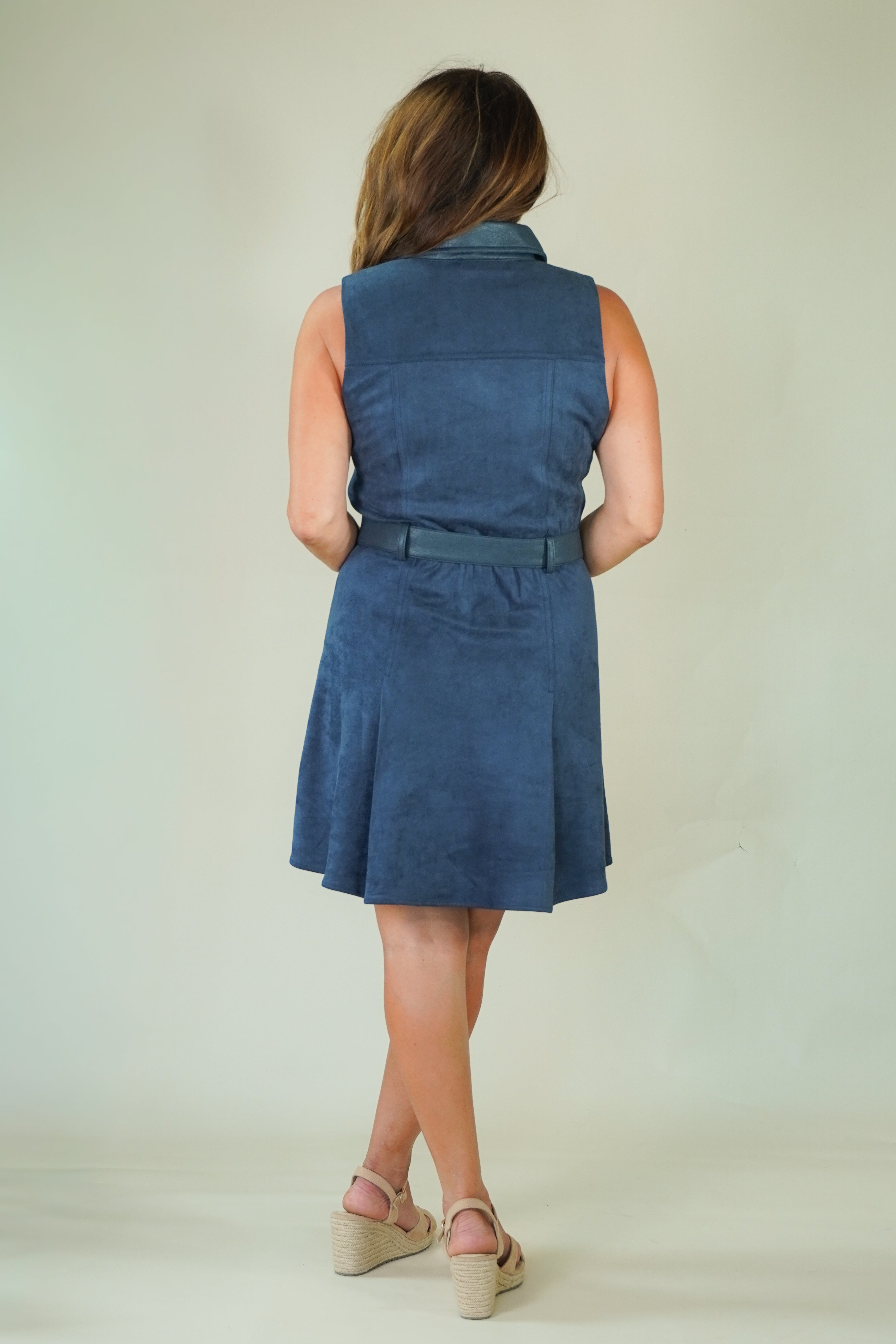 A woman viewed from behind wearing a sleeveless blue dress with a matching belt, standing against a plain light background.