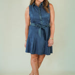 A woman modeling a sleeveless slate blue button-down shirt dress with a matching tie belt, paired with beige strappy sandals, against a plain light green studio background.