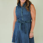 A woman modeling a sleeveless blue denim-like shirt dress with a button front, collared neckline, and matching tie belt, standing against a plain light beige studio background.