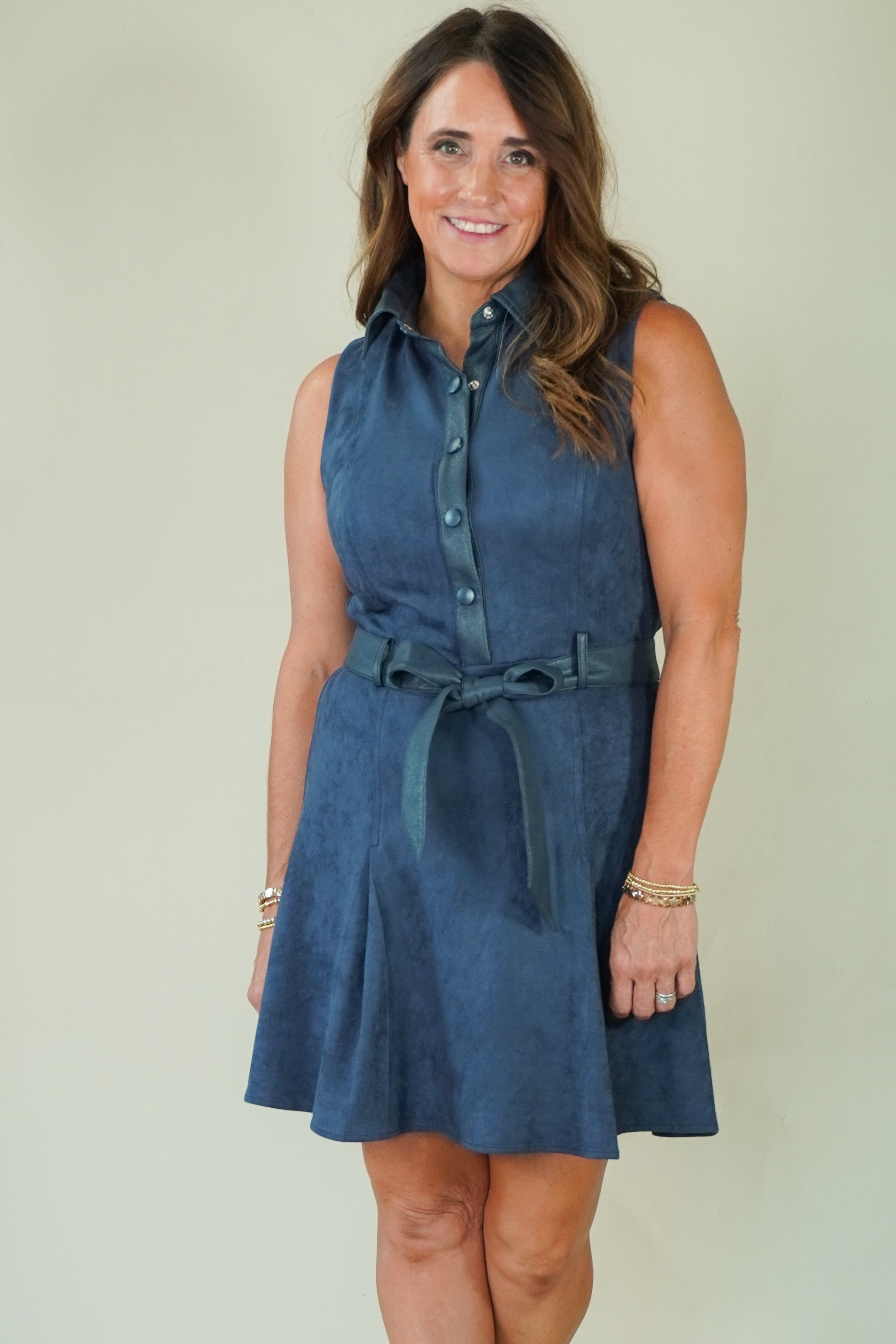 A woman modeling a sleeveless blue denim-like shirt dress with a button front, collared neckline, and matching tie belt, standing against a plain light beige studio background.