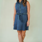 A woman modeling a sleeveless blue denim-like mini dress with a button-front neckline, tie belt at the waist, and a flared skirt, photographed against a plain light beige studio background.