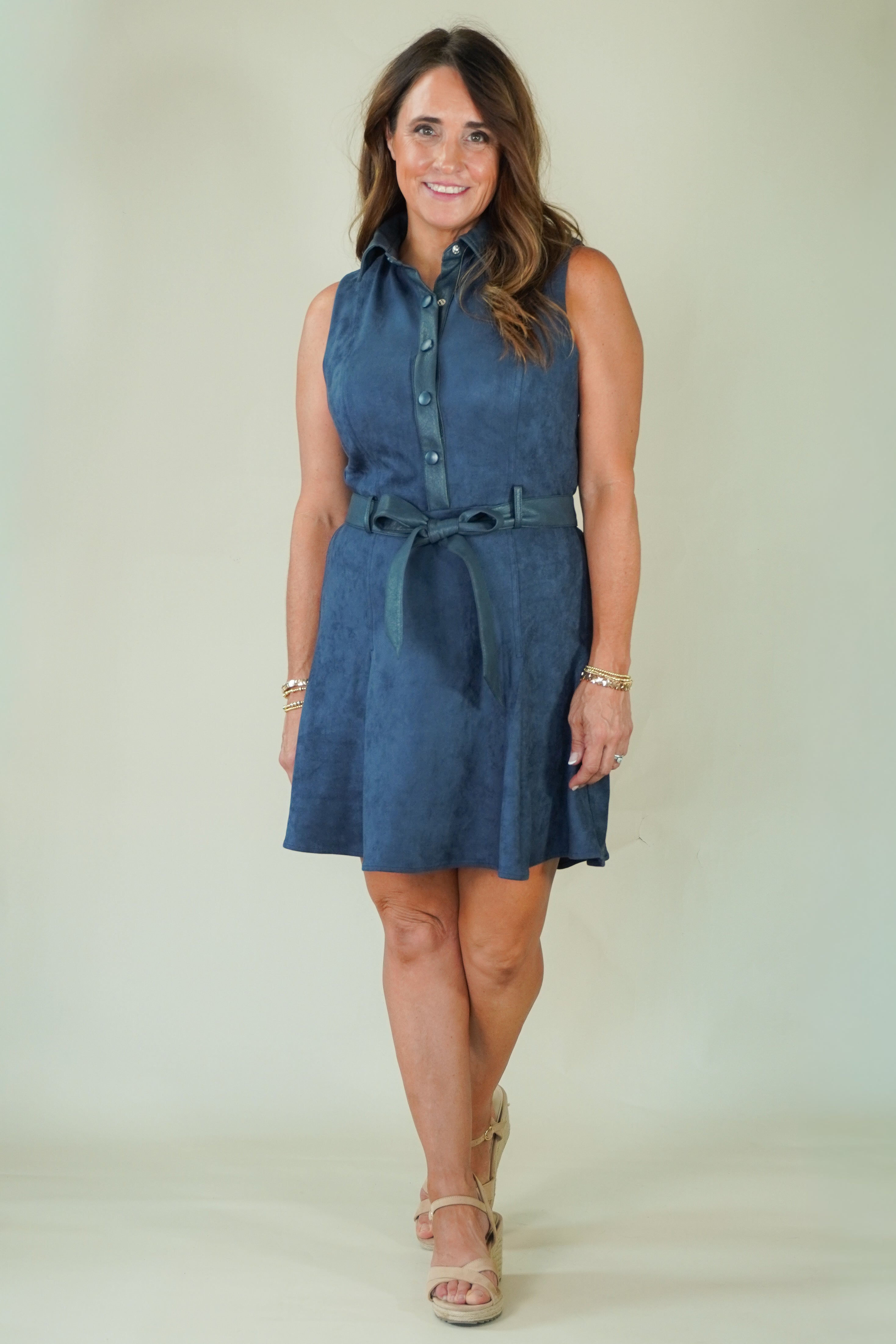 A woman modeling a sleeveless blue denim-like mini dress with a button-front neckline, tie belt at the waist, and a flared skirt, photographed against a plain light beige studio background.