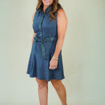 A woman modeling a sleeveless blue denim-like mini dress with a button-front neckline, tie waist, and flared skirt, standing against a plain light beige studio background.