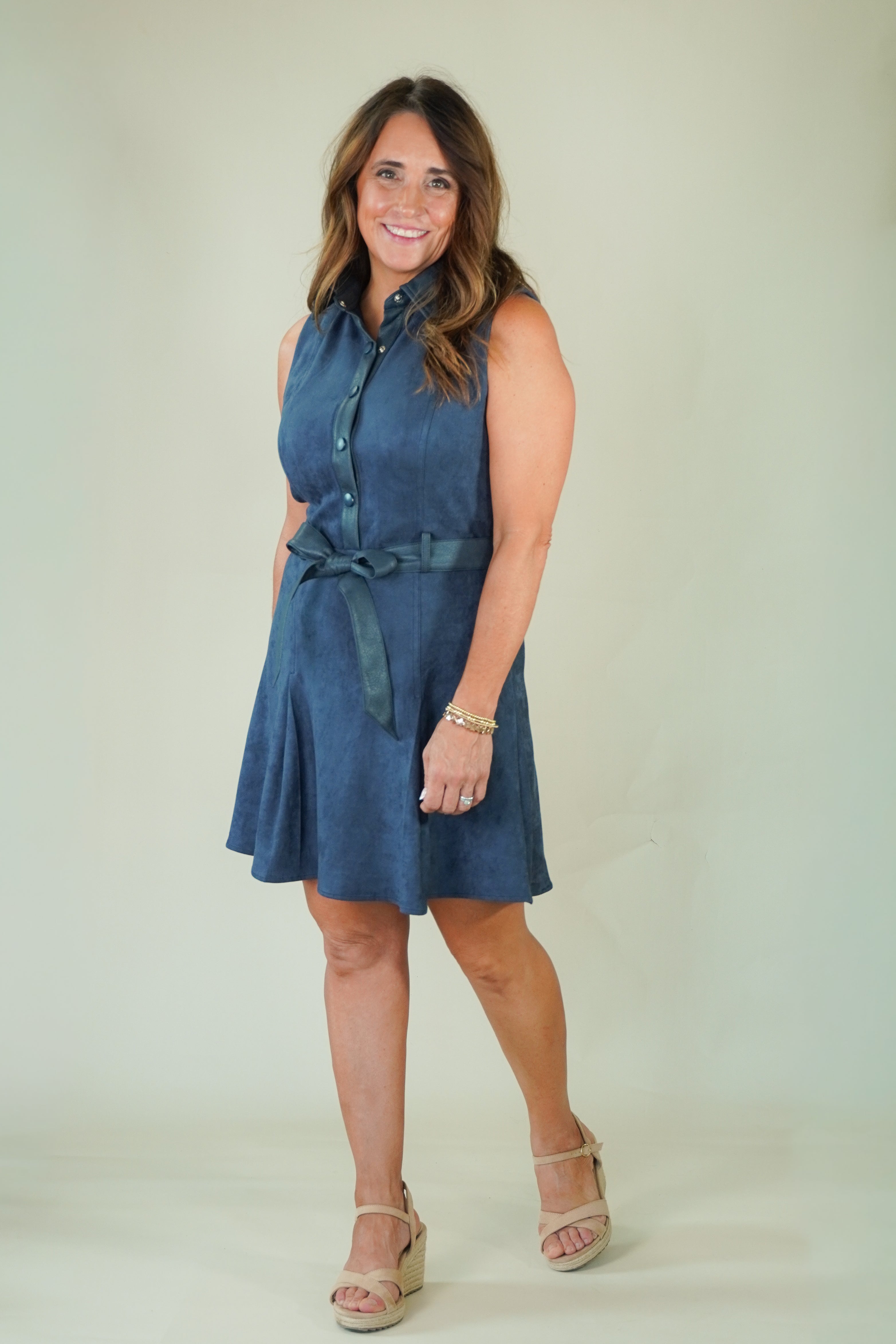 A woman modeling a sleeveless blue denim-like mini dress with a button-front neckline, tie waist, and flared skirt, standing against a plain light beige studio background.