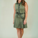 A woman modeling a sleeveless olive green shirt dress with a matching tie belt, standing against a plain light beige studio background.