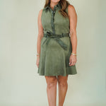 A woman modeling a sleeveless olive green dress with a buttoned collar, matching belt, and tie detail at the waist, paired with beige wedge sandals against a light beige studio background.