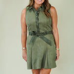 A woman modeling a sleeveless olive green mini dress with a button-up front, matching fabric tie belt, and beige strappy sandals, standing against a plain light beige studio background.