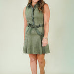 A woman modeling a sleeveless olive green mini dress with a buttoned collar, matching belt, and beige strappy heels against a light beige studio background.