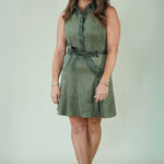 A woman modeling a sleeveless olive green dress with a button-up collar, matching belt, and beige strappy sandals against a light green studio backdrop.