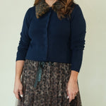 A woman modeling a navy blue cardigan with a brown and black leopard-print faux fur collar, paired with a matching leopard-print tulle midi skirt and black sandals, against a plain light beige studio background.
