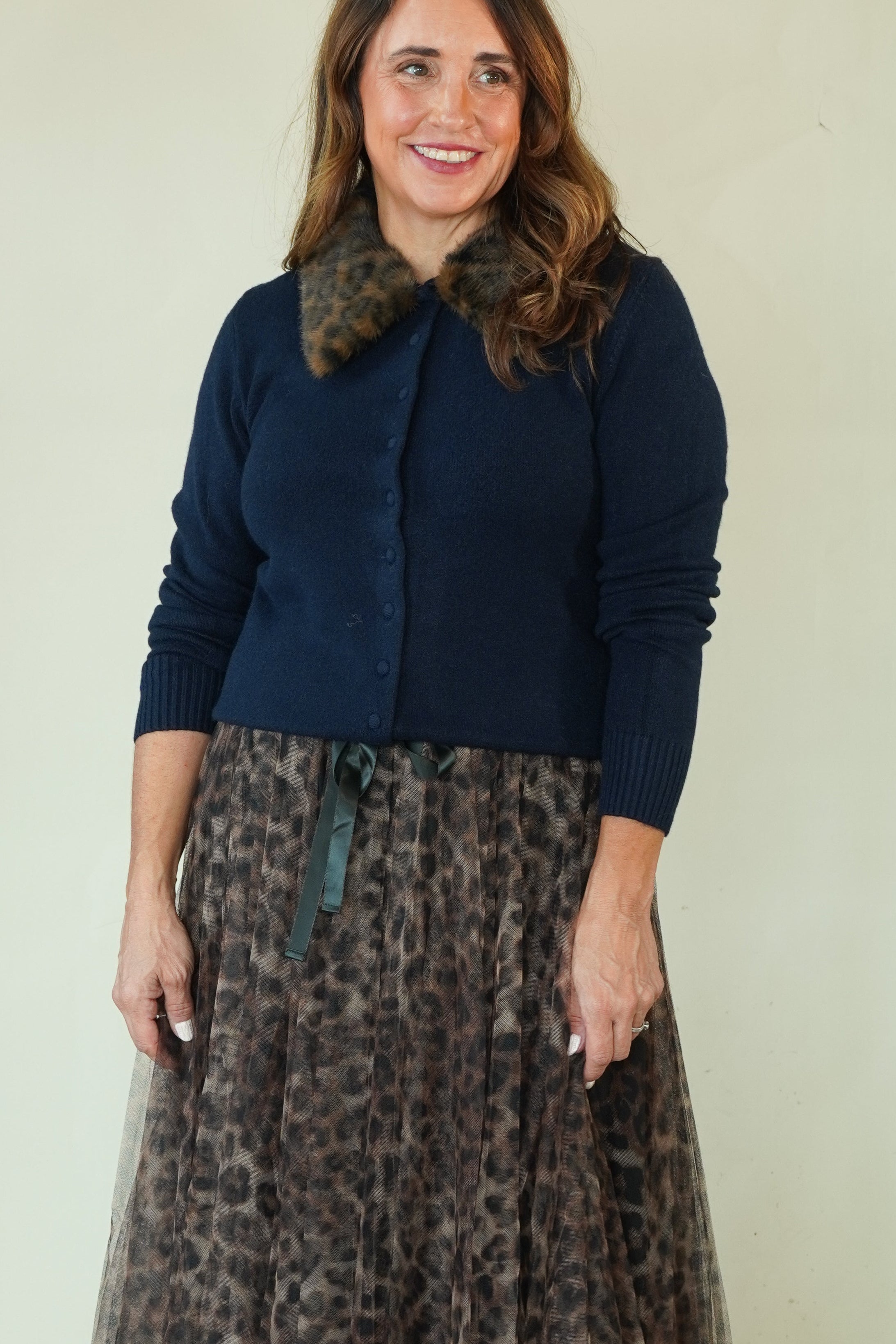 A woman modeling a navy blue cardigan with a brown and black leopard-print faux fur collar, paired with a matching leopard-print tulle midi skirt and black sandals, against a plain light beige studio background.