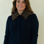 A woman modeling a navy blue button-up cardigan with a brown patterned collar, paired with white pants and brown shoes, standing against a plain off-white studio background.