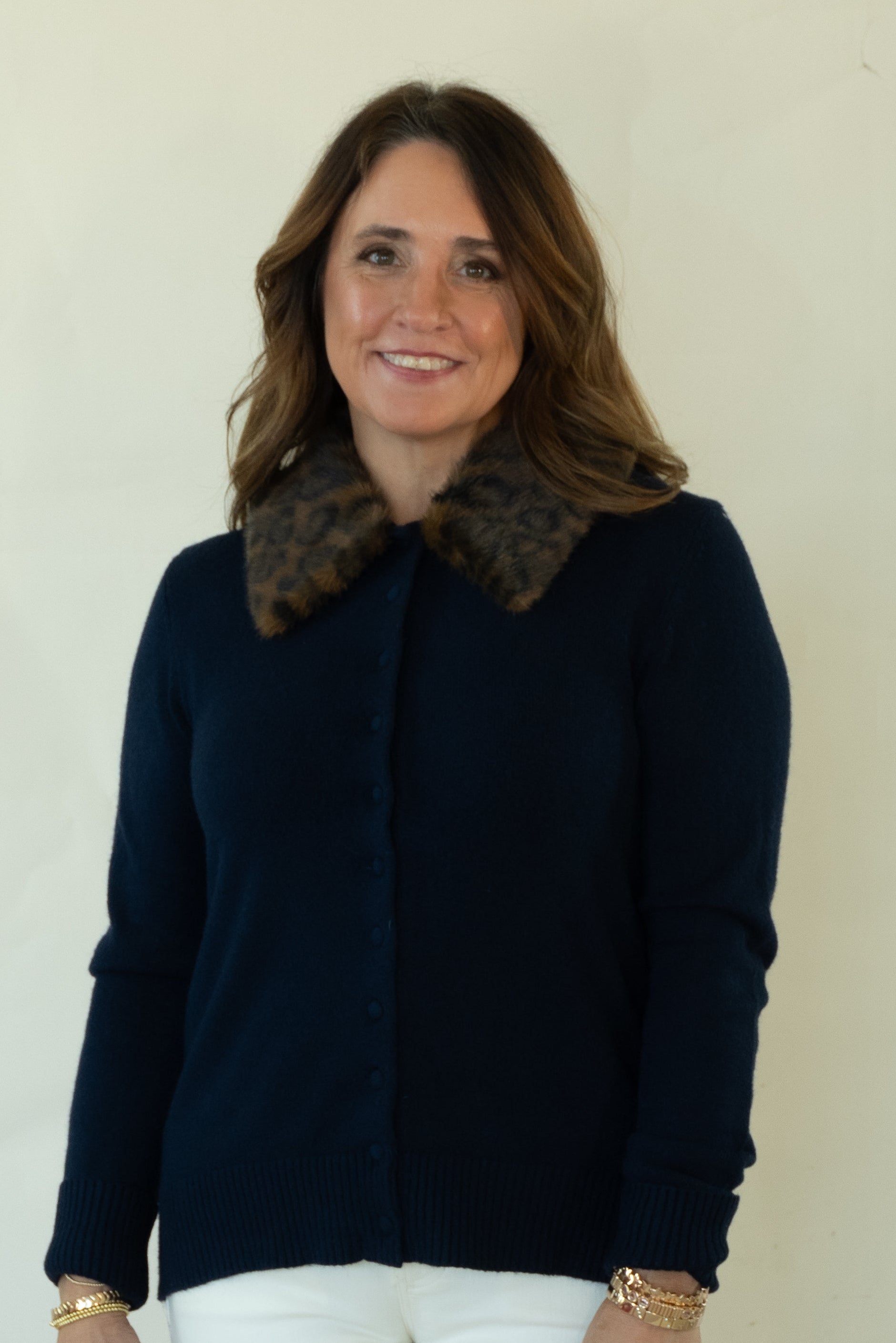 A woman modeling a navy blue button-up cardigan with a brown patterned collar, paired with white pants and brown shoes, standing against a plain off-white studio background.