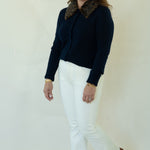 A woman modeling a dark navy blue knit cardigan with a brown patterned collar, paired with white pants and brown shoes, standing against a plain off-white studio background.