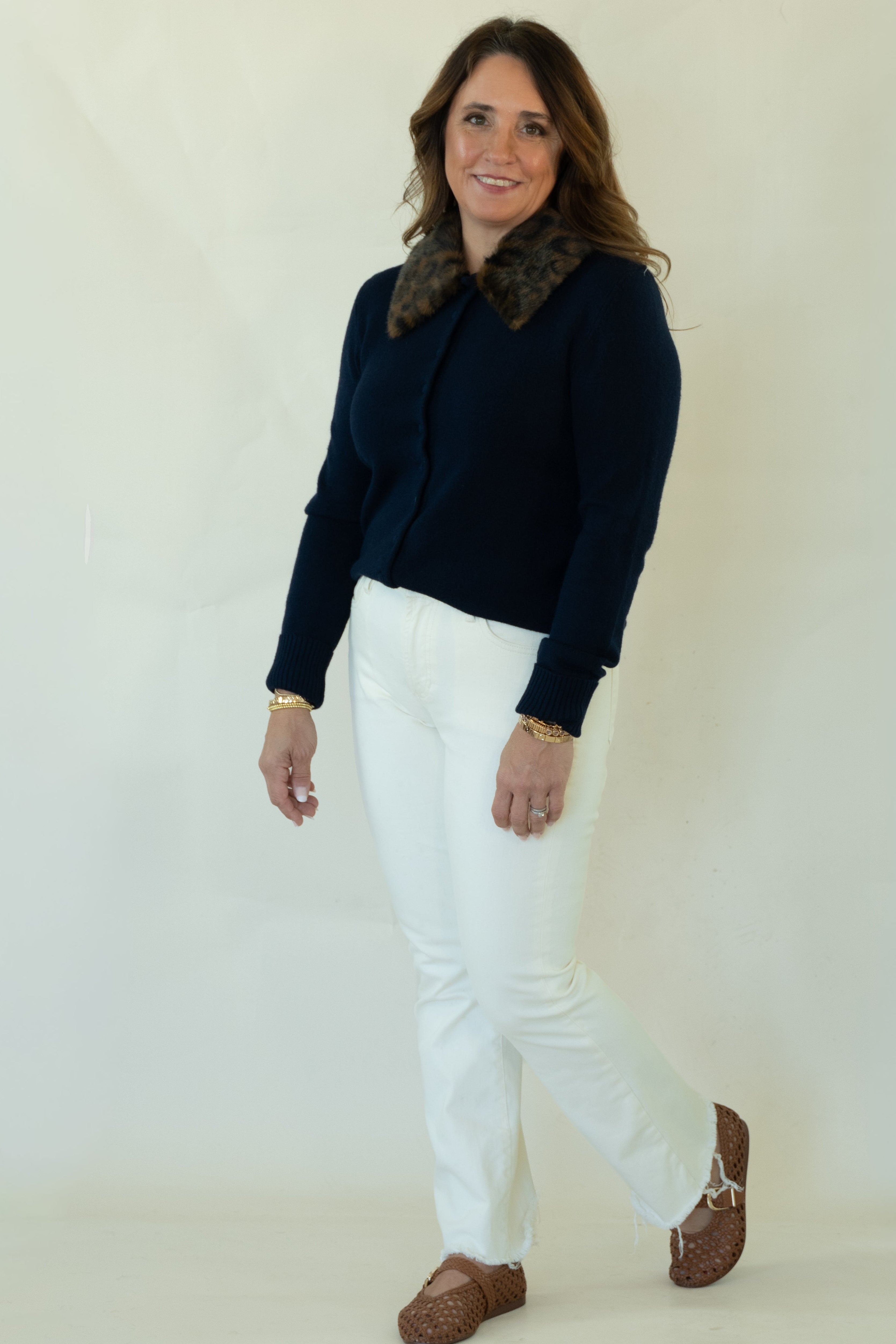 A woman modeling a dark navy blue knit cardigan with a brown patterned collar, paired with white pants and brown shoes, standing against a plain off-white studio background.