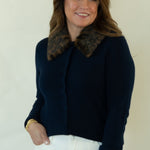 A woman modeling a dark navy blue knit cardigan with a brown patterned collar, paired with white pants and brown shoes, standing against a plain off-white studio background.