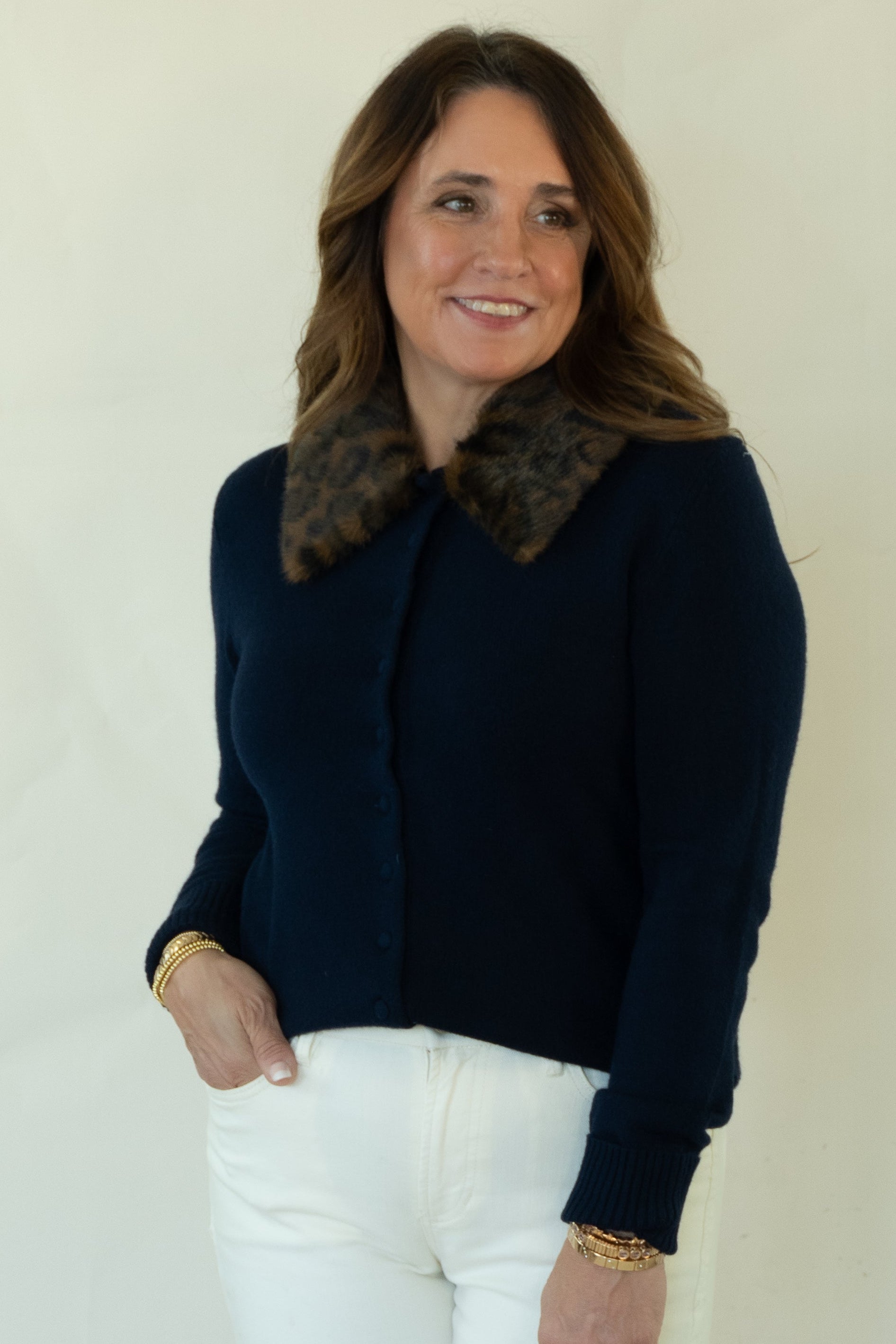 A woman modeling a dark navy blue knit cardigan with a brown patterned collar, paired with white pants and brown shoes, standing against a plain off-white studio background.