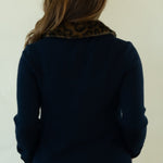 A woman shown from behind wearing a navy blue knit sweater with a brown patterned collar, white pants, and brown lace-up shoes, standing against a plain off-white background.