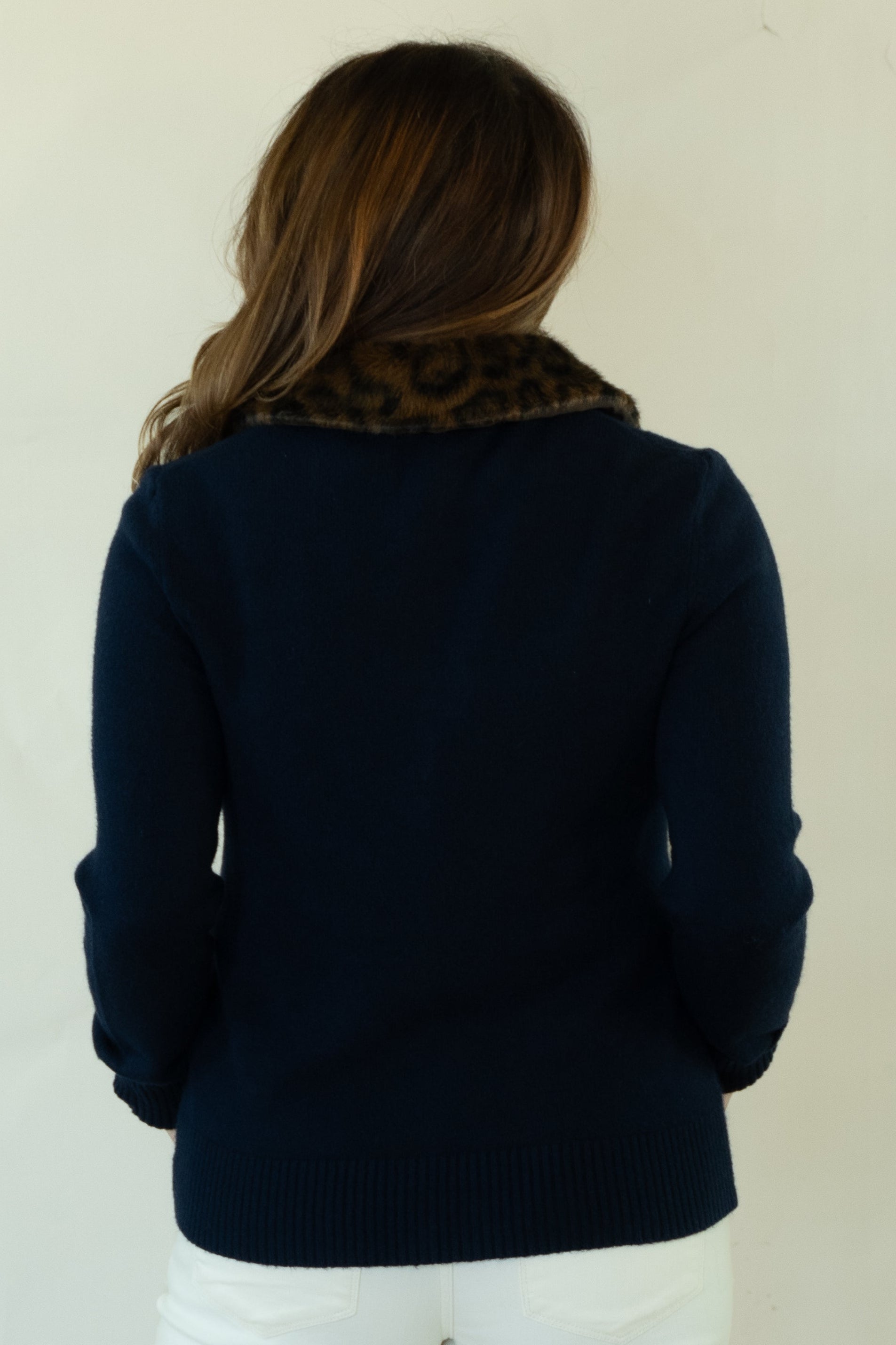 A woman shown from behind wearing a navy blue knit sweater with a brown patterned collar, white pants, and brown lace-up shoes, standing against a plain off-white background.