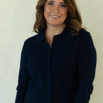 A woman modeling a dark navy blue zip-up cardigan sweater, white pants, and brown woven flat shoes, standing against a plain off-white studio background.