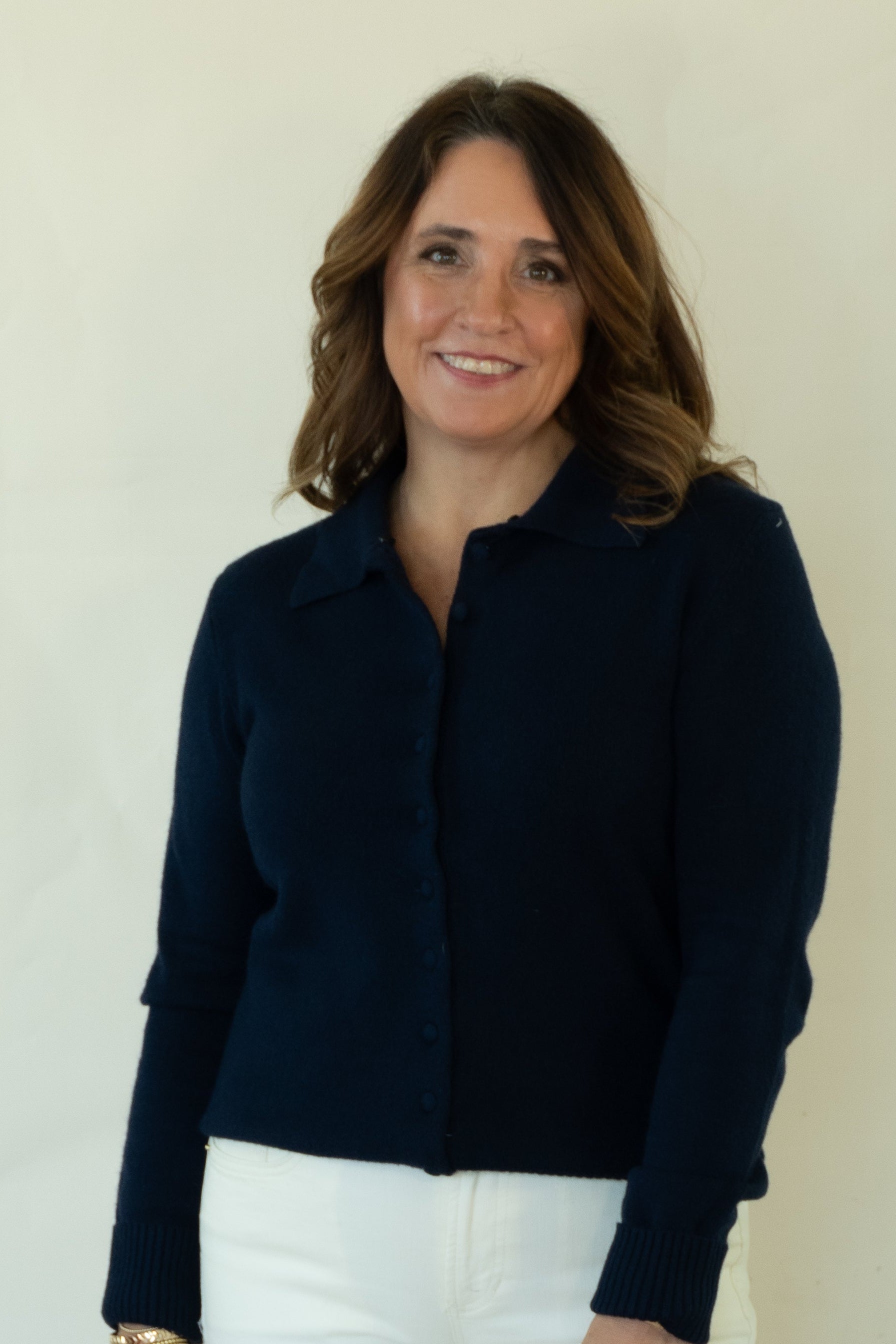 A woman modeling a dark navy blue zip-up cardigan sweater, white pants, and brown woven flat shoes, standing against a plain off-white studio background.