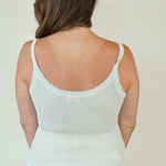 A woman viewed from the back wearing a light cream ribbed knit camisole top with thin straps and a scoop neckline, paired with white bottoms, against a plain light beige background.